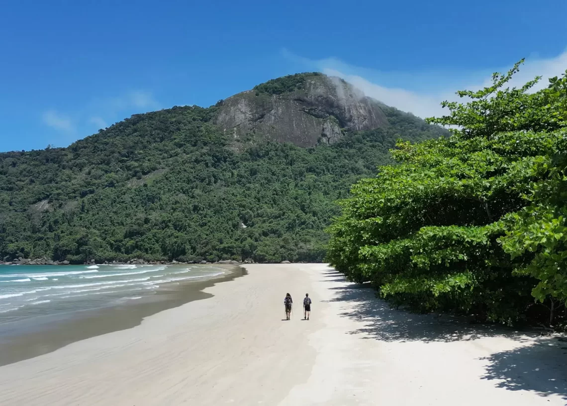 Two hikers walking on a wide, empty beach below a forested peak on Ilha Grande